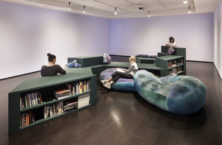 Modular bookshelf structure and large infinity shaped cushion in center of gallery with three figures seated reading books Modular bookshelf structure and large infinity shaped cushion in center of gallery with three figures seated reading books. Photo by Henrik Kam with additional editing by Brice Bischoff