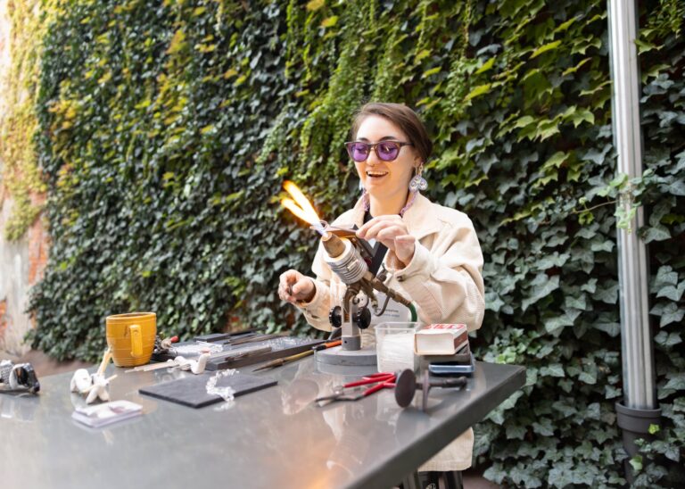 A female glass artist smiles while working on a small project over an open flame. She is wearing purple protective glasses and sitting in front of a wall covered in ivy. A female glass artist smiles while working on a small project over an open flame. She is wearing purple protective glasses and sitting in front of a wall covered in ivy.