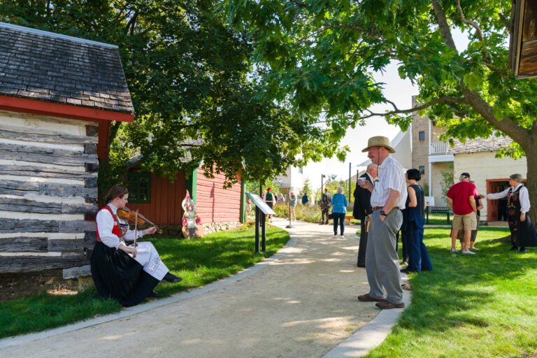 Visitors to Vesterheim listening to musician Eden Ehm playing the Hardanger Fiddle, Norway's national instrument, in Vesterheim Heritage Park. Vesterheim