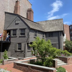 A gray clapboarded two-story 17th century home with an overhanging second floor situated next to a tall building on one side and a brick courtyard with garden beds and a pear tree on the other side. A second floor walkway provides access to the second floor and is hung with red, white, and blue bunting. The Paul Revere Memorial Association