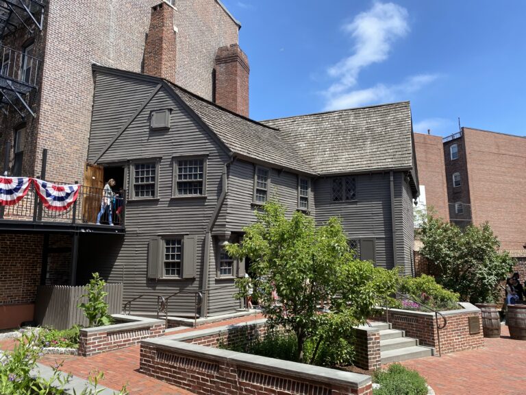 A gray clapboarded two-story 17th century home with an overhanging second floor situated next to a tall building on one side and a brick courtyard with garden beds and a pear tree on the other side. A second floor walkway provides access to the second floor and is hung with red, white, and blue bunting. The Paul Revere Memorial Association