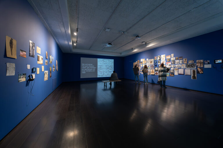 Figures standing and sitting in blue gallery featuring projected video on one end and archival imagery arranged across the adjacent walls Figures standing and sitting in blue gallery featuring projected video on one end and archival imagery arranged across the adjacent walls. Photo by Adam Hays