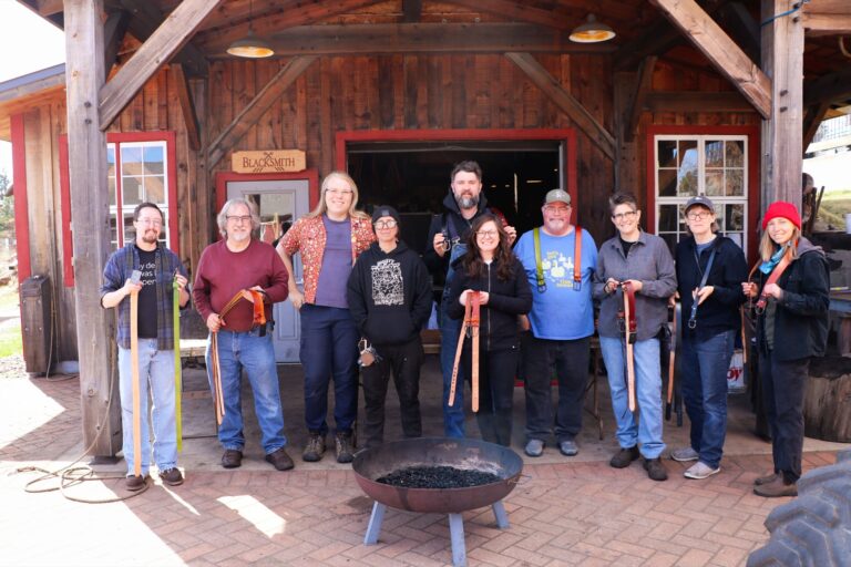 A North House belt-making class standing outside the Blacksmith Shop on campus with their finished leather and metal hand-made belts. North House Folk School