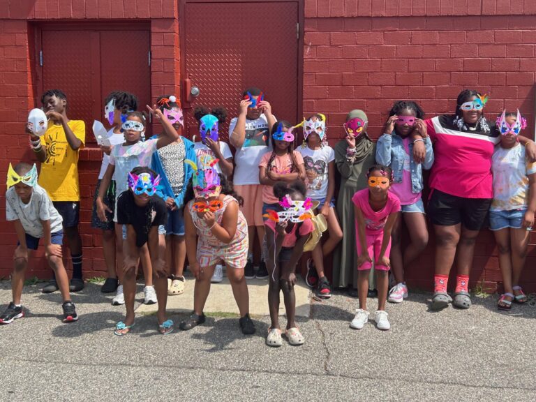 a group of children wearing colorful masks pose in front of a red brick wall a group of children wearing colorful masks pose in front of a red brick wall