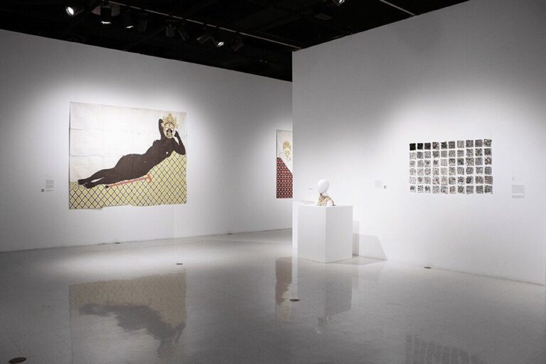 Gallery installation image with three artworks. There is one large tapestry depicting a nude reclining female figure with a walking cane, one cream colored wool scarf on a mannequin bust and a grid of black and white wool squares. Installation view: Script/Rescript, University Art Gallery; Image courtesy San Diego State University School of Art and Design