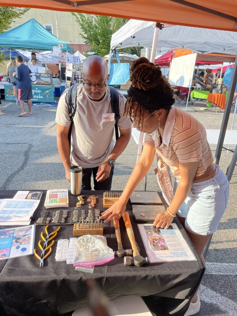 A young woman with dreadlocks is explaining how to stamp metal to a mid-aged man that shows interest in her booth at the farmers market. Molly Shulman A young woman with dreadlocks is explaining how to stamp metal to a mid-aged man that shows interest in her booth at the farmers market. Molly Shulman