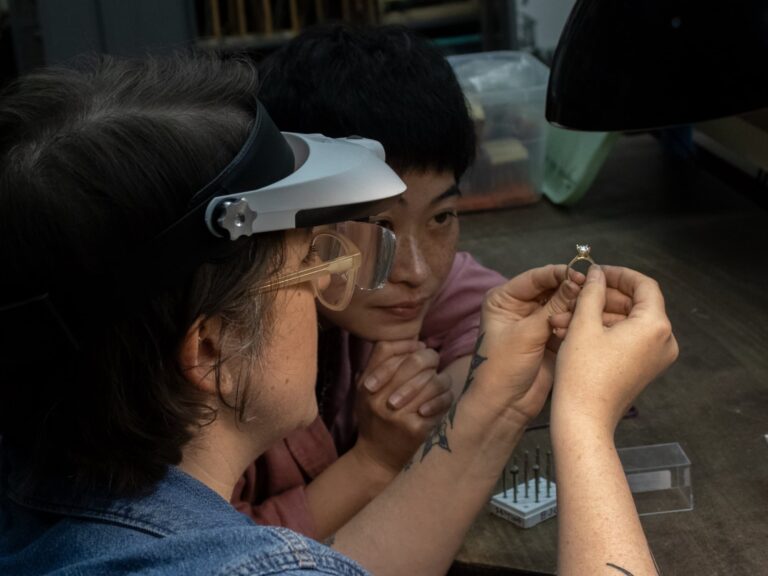 Two women closely examing a prong setting for a gem stone. J Taran Diamond Two women closely examing a prong setting for a gem stone. J Taran Diamond