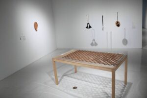 Gallery installation image of five individual artworks. There is one ceramic artwork on the wall in the shape of a nopales cactus pad. There are three hanging leather and metal sculptures shaped like a bell, a pocket knife and a tea pot. There is a large wooden table in the foreground with a clay chainlink fence on its surface. Installation view: Crossing the Line, University Art Gallery; Image courtesy San Diego State University School of Art and Design