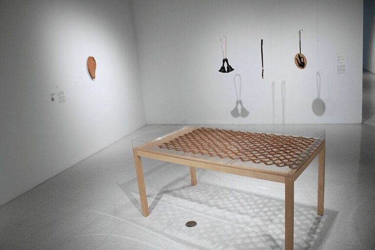 Gallery installation image of five individual artworks. There is one ceramic artwork on the wall in the shape of a nopales cactus pad. There are three hanging leather and metal sculptures shaped like a bell, a pocket knife and a tea pot. There is a large wooden table in the foreground with a clay chainlink fence on its surface. Installation view: Crossing the Line, University Art Gallery; Image courtesy San Diego State University School of Art and Design