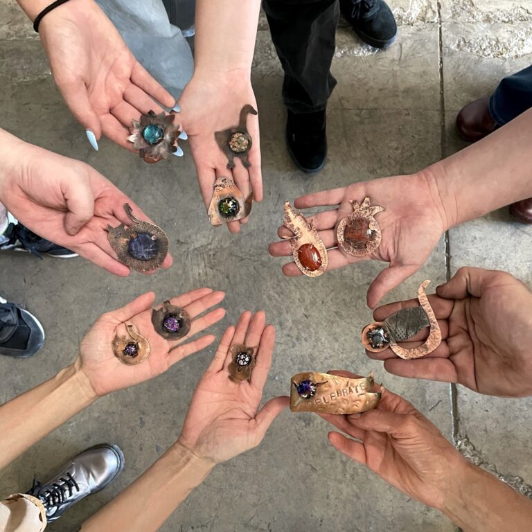 A circle of hands holding copper jewelry with colorful glass cabochons set in them. Adam's Forge