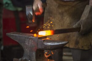 Close up of a yellow-hot piece of steel on an anvil being hit by a hammer. Hoot scale is flying off. The smith is in a leather apron and a leather glove on their stock hand. Martha Benedict