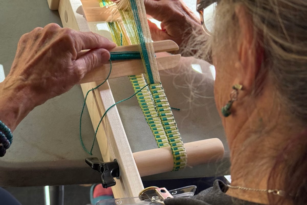 A person is shown from the side, intently weaving a colorful patterned band on a small loom. American Swedish Institute