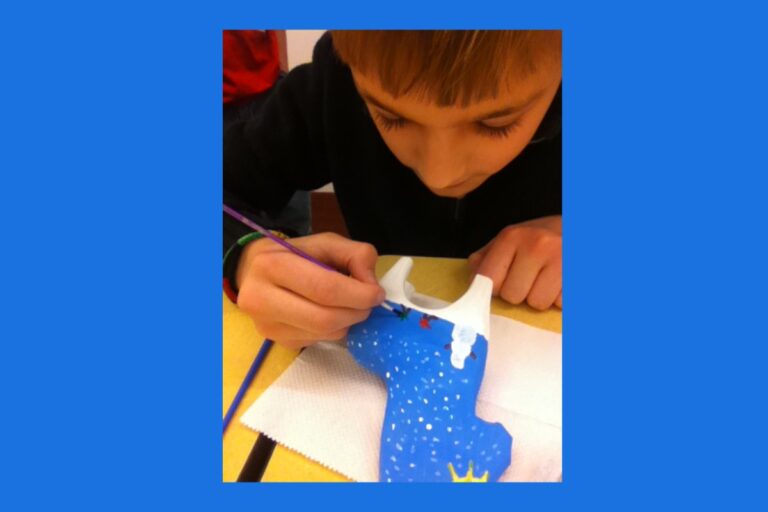 A young boy is carefully painting a blue ceramic object, adding white dots and other details. He is focused on his artwork with a brush in his hand. American Swedish Institute