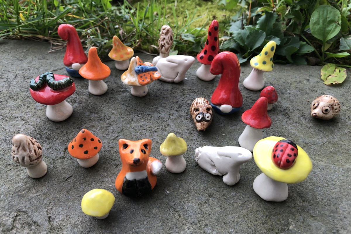 A collection of small, handmade ceramic figures, including mushrooms, gnomes, hedgehogs, a fox, and a caterpillar, are arranged on a stone surface with grass in the background. American Swedish Institute