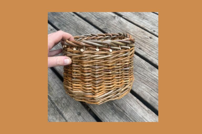 A hand holds a small, handmade wicker basket against a background of weathered wooden planks. The basket is woven with natural materials. American Swedish Institute