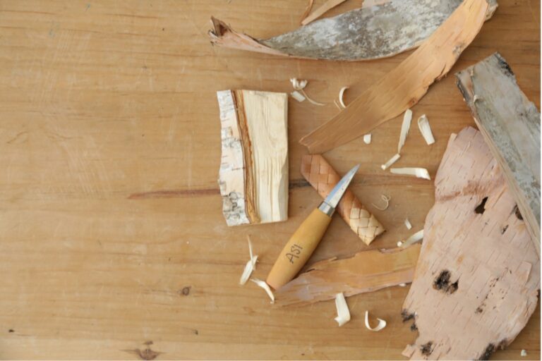 A wooden workbench displays birch bark pieces, shavings, and a small knife with a wooden handle. American Swedish Institute