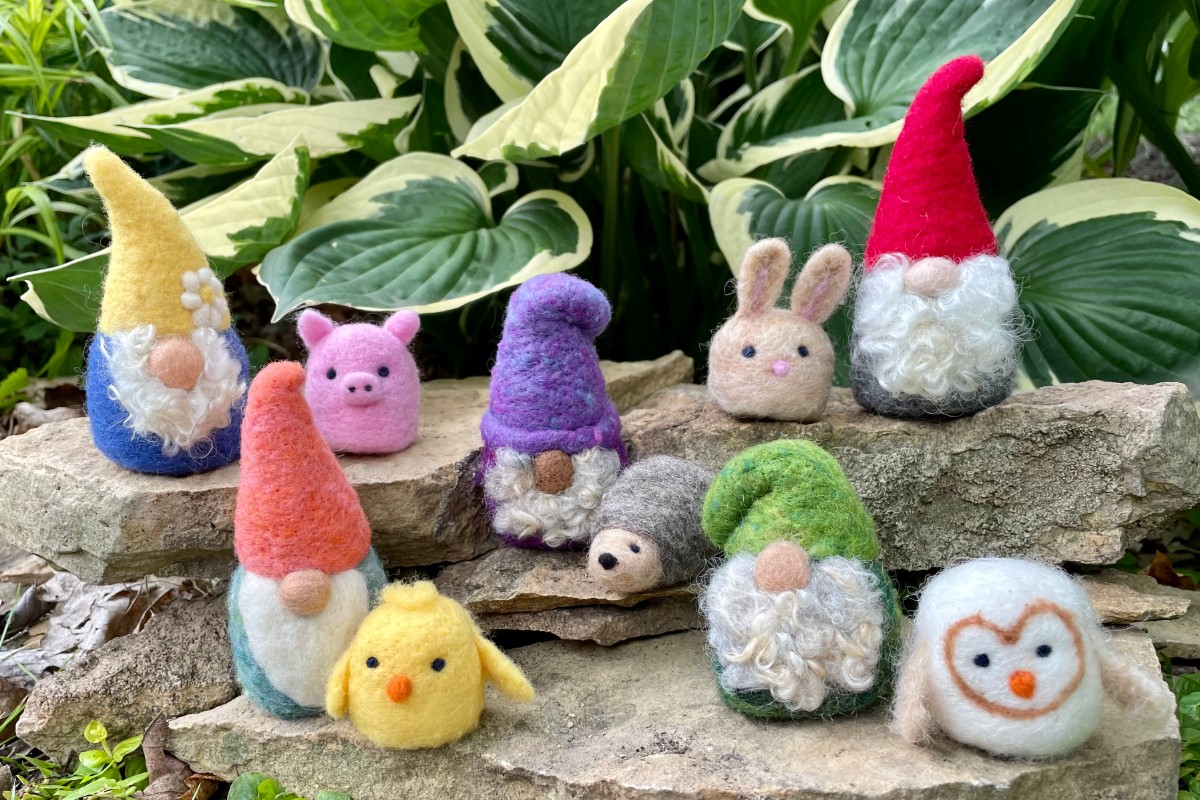 A collection of small, felted gnomes and animal figures are displayed on rocks in front of lush green foliage. American Swedish Institute
