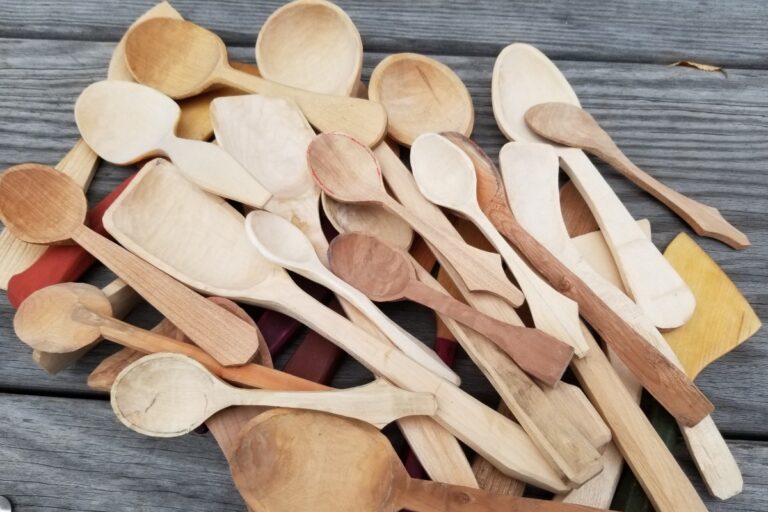 A collection of handmade wooden spoons of various sizes and wood types are piled on a weathered wooden surface. The spoons are carved and sanded, showcasing different wood grains and shapes. American Swedish Institute
