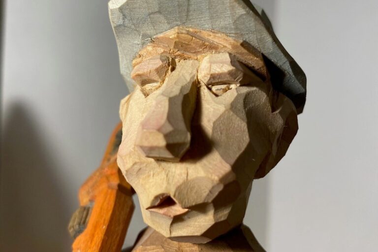 A close-up shot of a wooden carving of a person's face wearing a hat. The carving has a rough, textured finish with visible chisel marks. American Swedish Institute