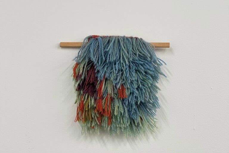 A textured, multi-colored fiber art piece hangs from a wooden dowel against a white wall. American Swedish Institute