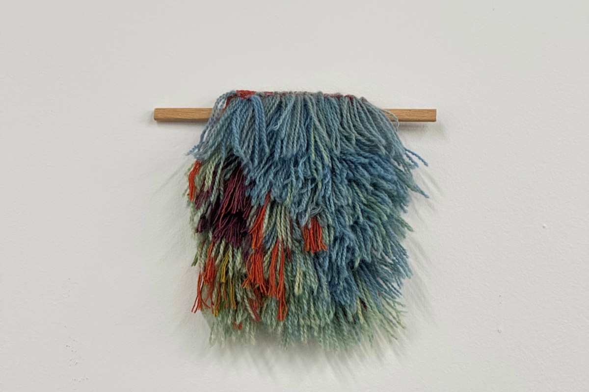 A textured, multi-colored fiber art piece hangs from a wooden dowel against a white wall. American Swedish Institute