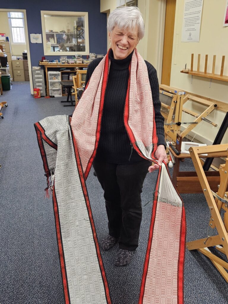 Artisan Joanna is standing next to her loom, wrapped in her completed warp. This fabric will be finished into scarves. Hartford Artisans Weaving Center