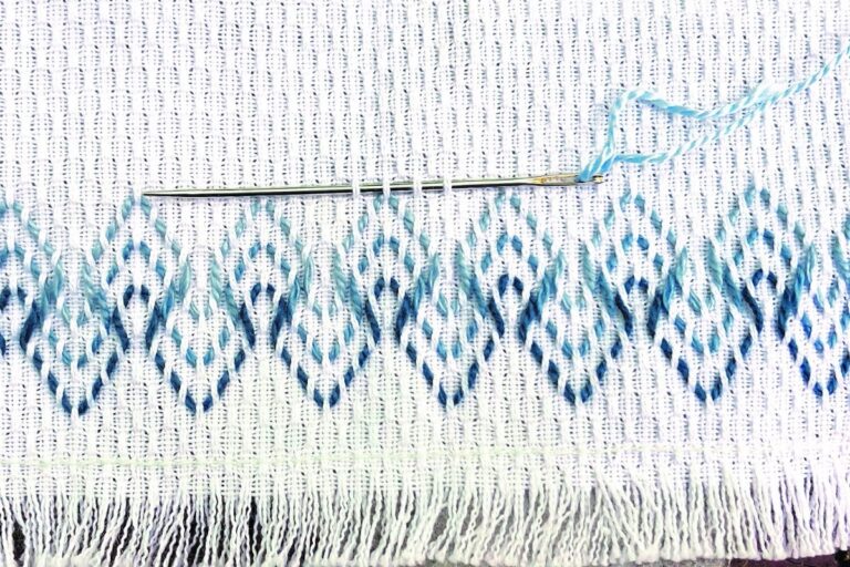 A needle stitches blue yarn on white fabric. American Swedish Institute