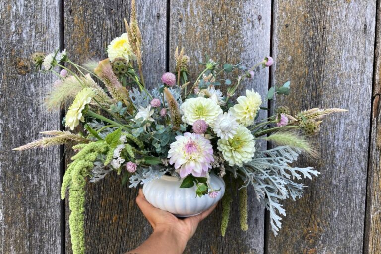 A hand holds a vase of flowers against a wooden background. American Swedish Institute