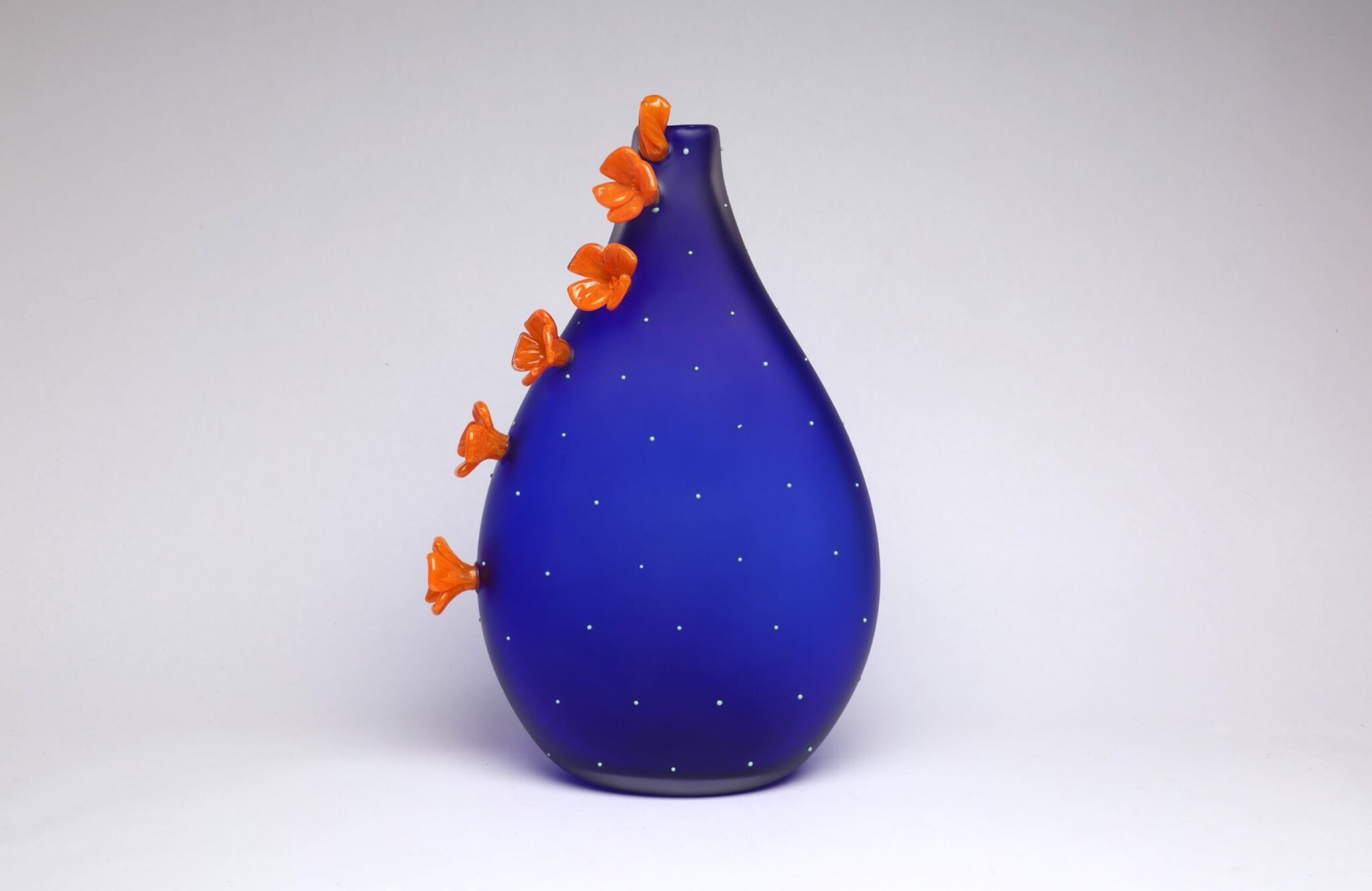 This is a photo of the large California Poppy Vessel that was created in the video. It is a bright, matte, cobalt-colored body with mint dots and vibrant orange and yellow poppy flowers that trail the side of the Vessel. Photo by Hannah Nicholson of Nicholson van Altena Glass