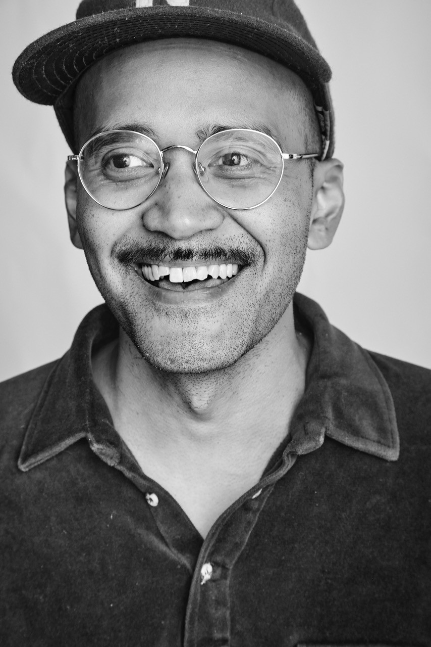 Charles Thompson. Close up of a smiling face of a person wearing a baseball cap and glasses. The image is black and white and their mustache is dark