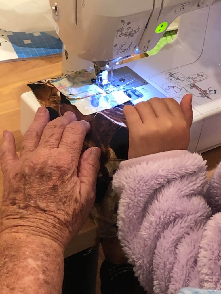 An elder teaches a young child to quilt for the first time. Their hands work together at the sewing machine, part of a tradition open to makers ages 4 to 104 An elder teaches a young child to quilt for the first time. Their hands work together at the sewing machine, part of a tradition open to makers ages 4 to 104. Image by Jayna Zweiman